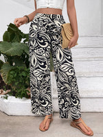 Person wearing black and white patterned wide-leg pants with a white top and sandals, standing next to a plant.