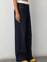 A person wearing dark indigo wide-leg jeans with a white cropped top and yellow slip-on shoes, standing against a plain light gray wall.