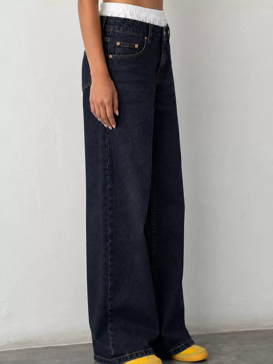 A person wearing dark indigo wide-leg jeans with a white cropped top and yellow slip-on shoes, standing against a plain light gray wall.