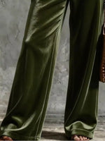 Olive green wide-leg velvet pants shown on a model, featuring a satin-like sheen and draped fabric folds, paired with brown sandals against a textured gray background.
