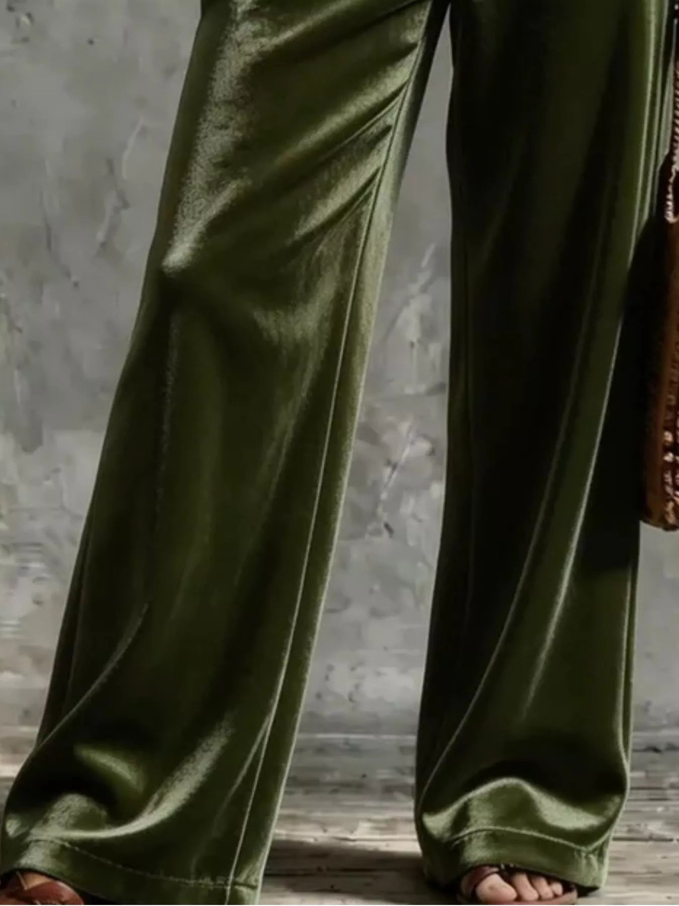 Olive green wide-leg velvet pants shown on a model, featuring a satin-like sheen and draped fabric folds, paired with brown sandals against a textured gray background.