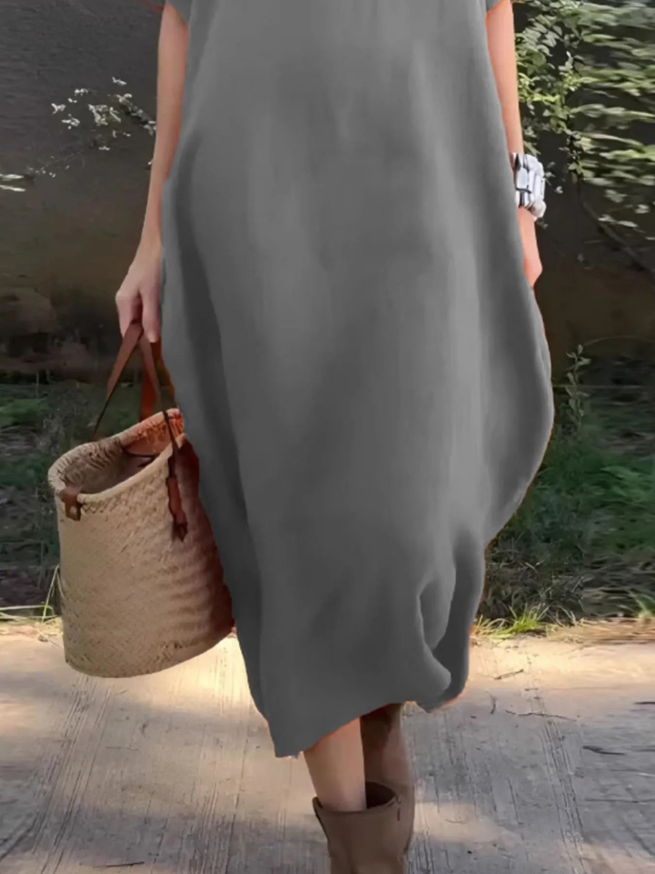Person wearing a gray dress holding a woven bag by a body of water.