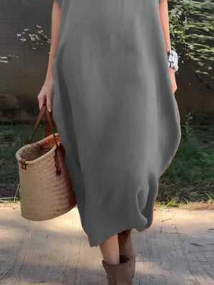 Person wearing a gray dress holding a woven bag by a body of water.