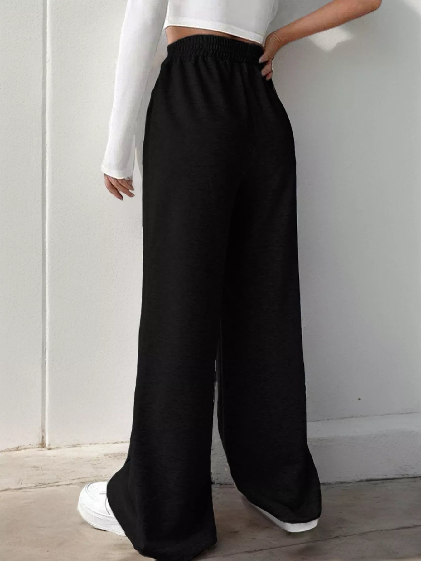 Person wearing black wide-leg pants with a white top against a light gray wall.