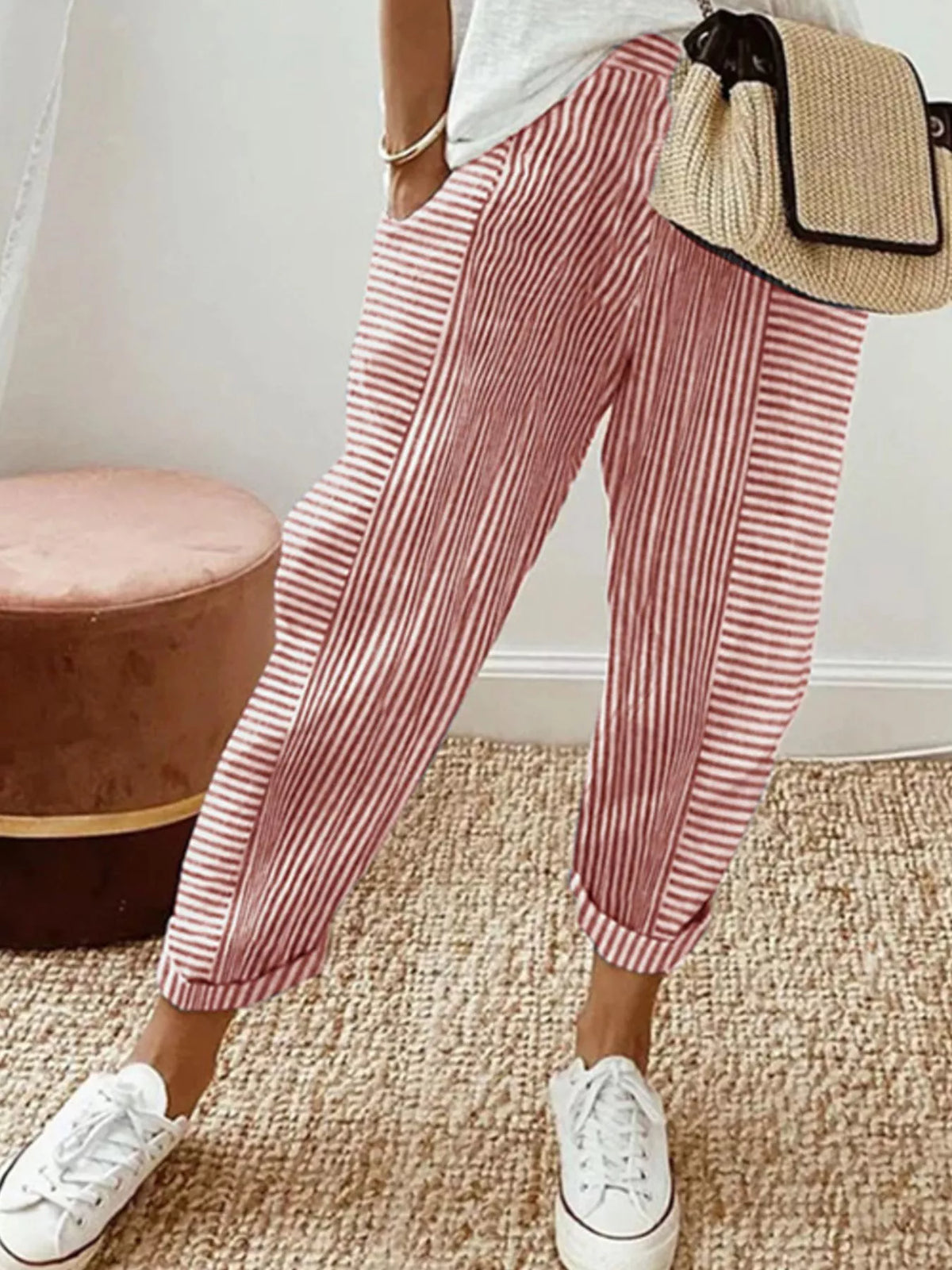 A person wearing loose, cuffed pink and white vertically striped pants, white sneakers, and carrying a woven straw shoulder bag in an indoor setting.