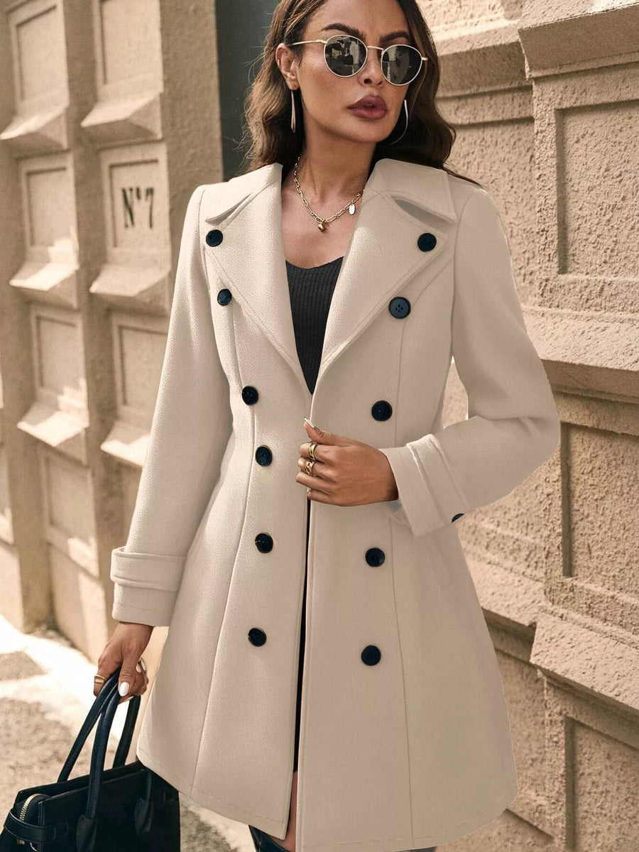 Woman wearing a beige coat with black buttons against a textured wall.