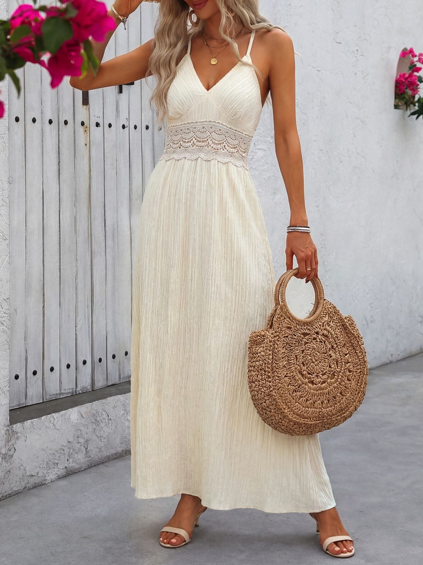 Woman in a white dress holding a woven bag with a white wall and pink flowers in the background