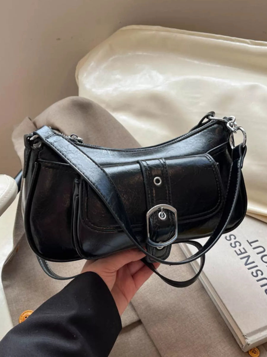A black faux-leather crescent-shaped shoulder bag with a silver buckle detail and a long strap, held by a hand over a beige fabric surface with a cream-colored bag in the background.