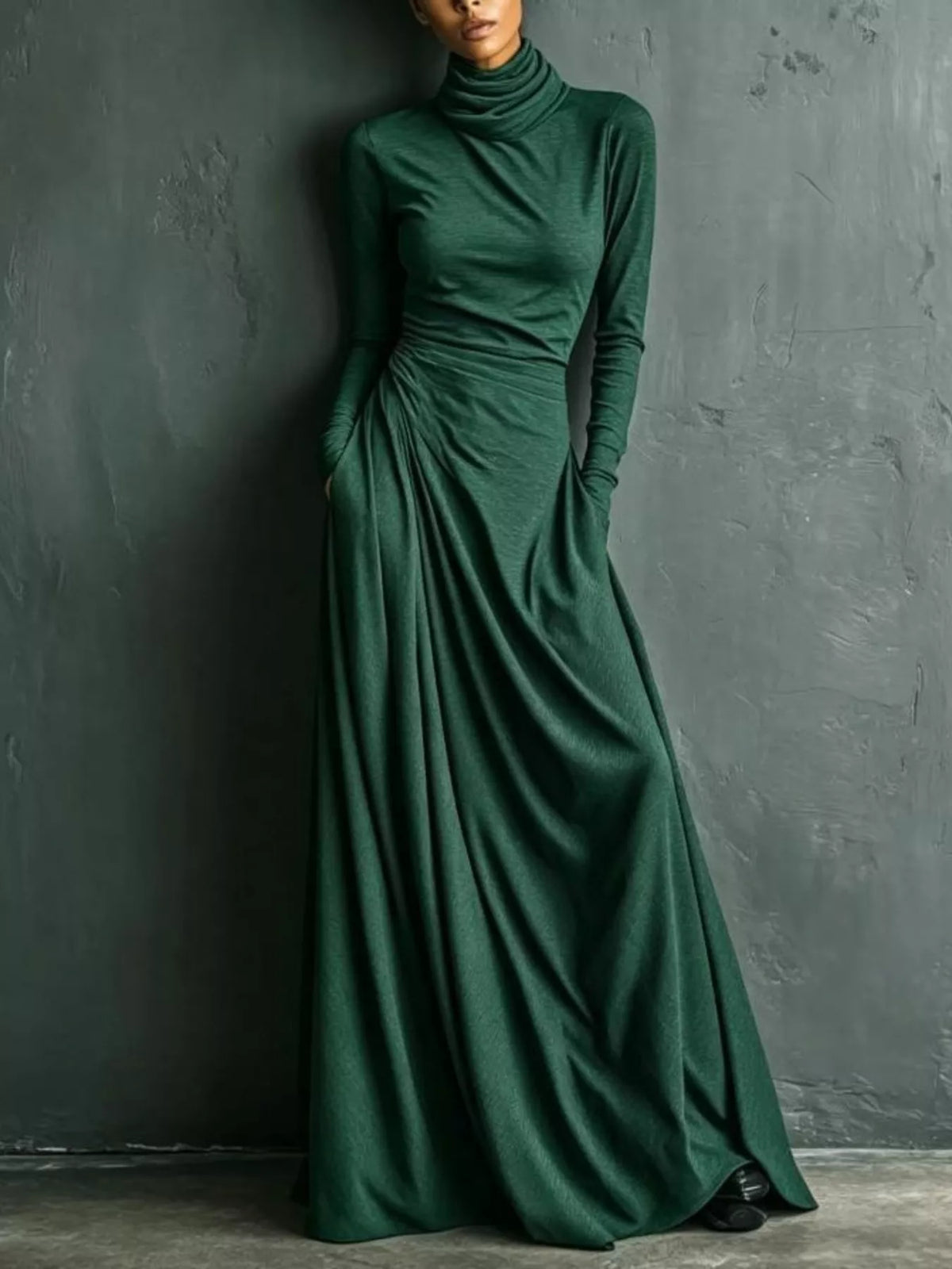 A woman wearing a long-sleeve, high-neck, floor-length dark green dress with gathered draping at the waist, standing against a textured dark gray wall.