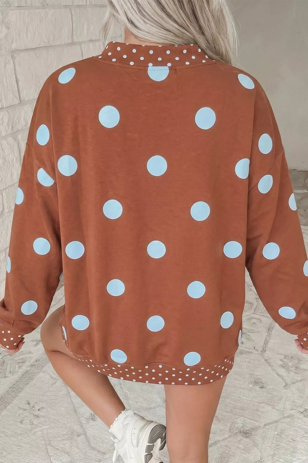 A person wearing a rust-orange long-sleeve sweatshirt with large light blue polka dots, shown from the back in an indoor setting with a light brick wall background.