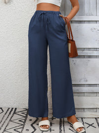Person wearing blue wide-leg pants with a white top and sandals, holding a brown handbag against a textured wall.