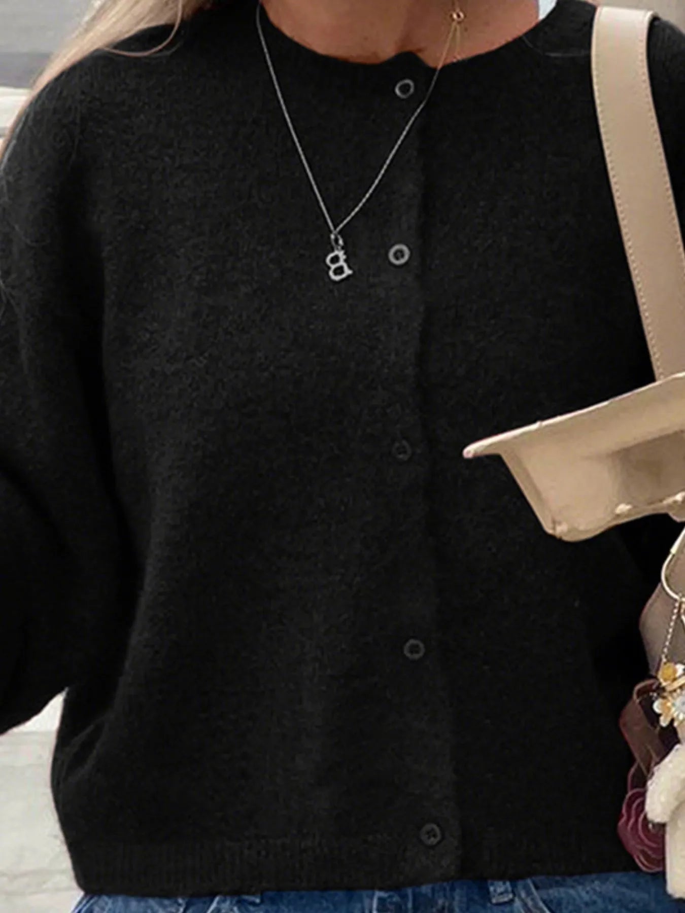 Person wearing a black cardigan with a necklace, holding a beige handbag.