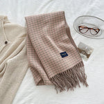 Plaid scarf with tassels on a white surface next to a beige cardigan and glasses.