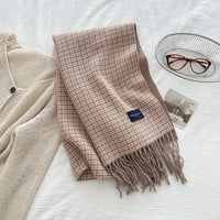 Plaid scarf with tassels on a white surface next to a beige cardigan and glasses.