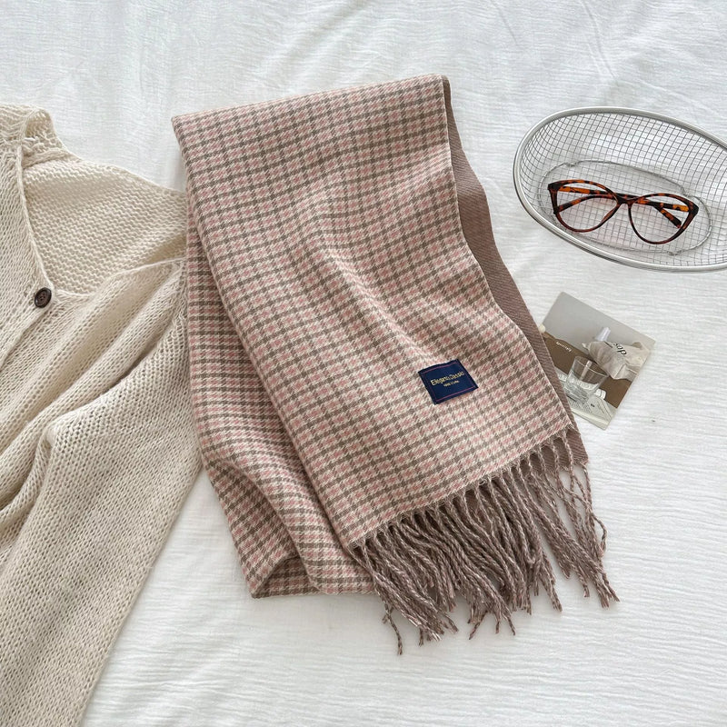 Plaid scarf with tassels on a white surface next to a beige cardigan and glasses.