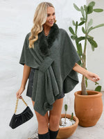 Woman wearing a dark green poncho with fur trim, standing next to a potted plant.