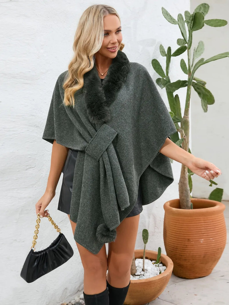 Woman wearing a dark green poncho with fur trim, standing next to a potted plant.