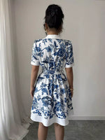 Person wearing a white dress with blue floral patterns, standing against a plain wall.