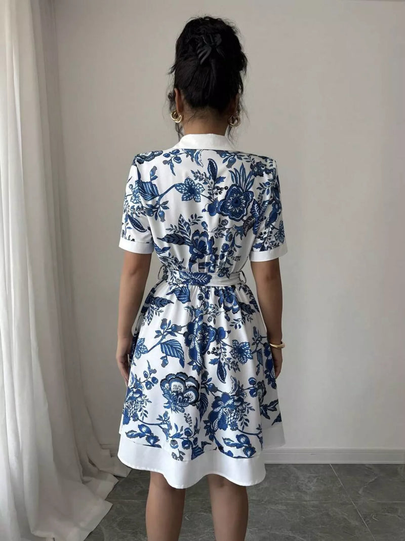 Person wearing a white dress with blue floral patterns, standing against a plain wall.