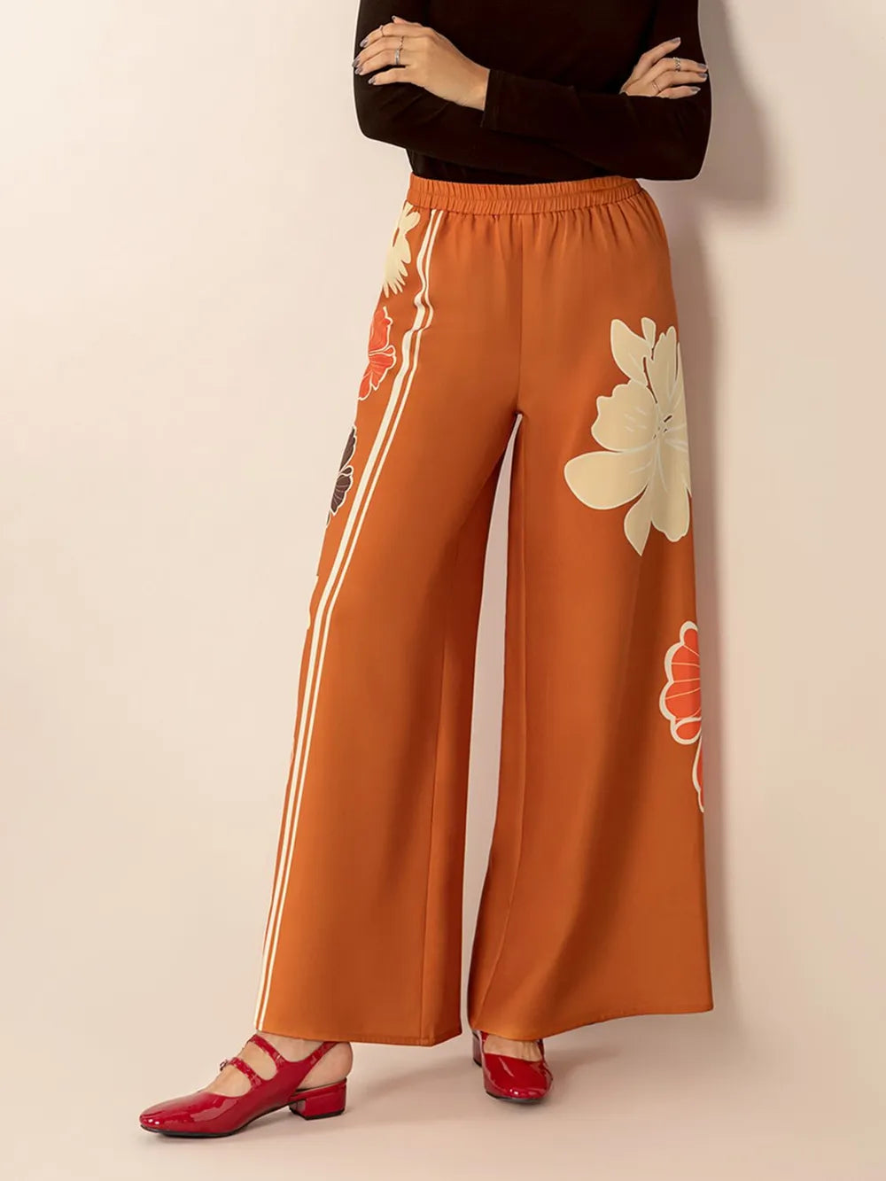 Orange wide-leg pants with floral designs worn by a person on a beige background