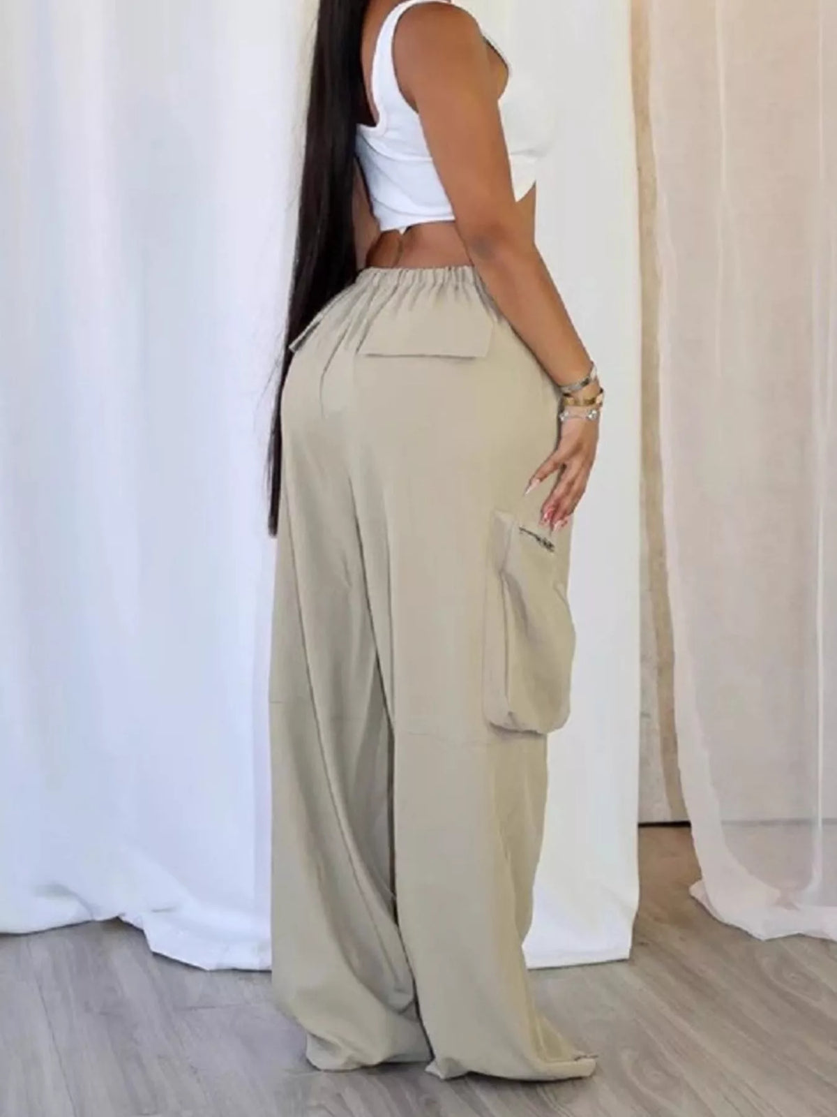 A woman wearing loose, wide-leg beige cargo pants with a white crop top, standing in front of white draped curtains.