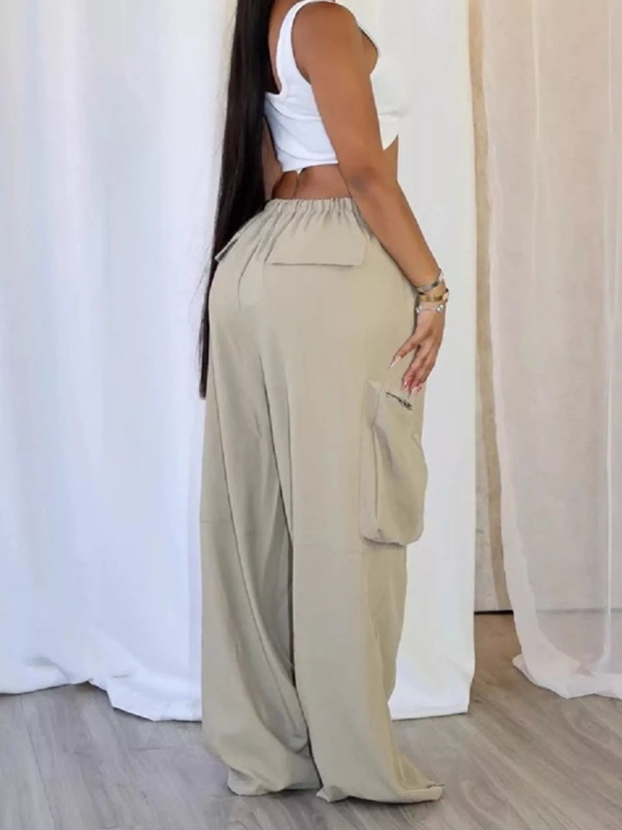 A woman wearing loose, wide-leg beige cargo pants with a white crop top, standing in front of white draped curtains.