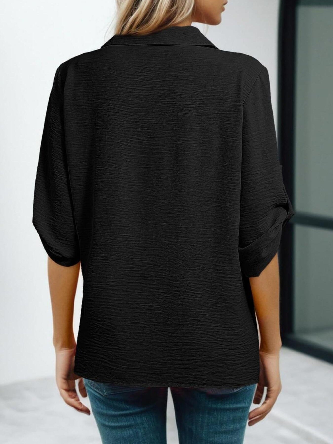 Person wearing a black textured blouse with rolled-up sleeves and blue jeans on a light background