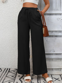 Person wearing black wide-leg pants with a white top and sandals, standing against a wooden panel wall.