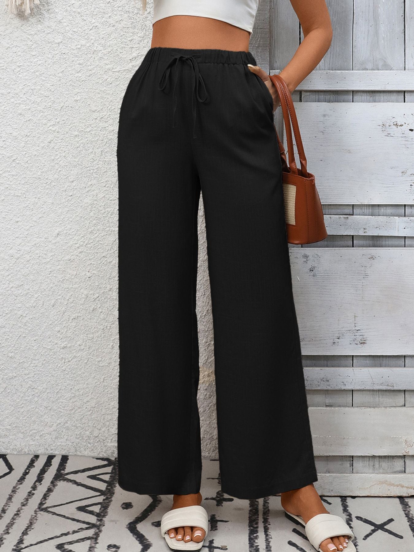 Person wearing black wide-leg pants with a white top and sandals, standing against a wooden panel wall.