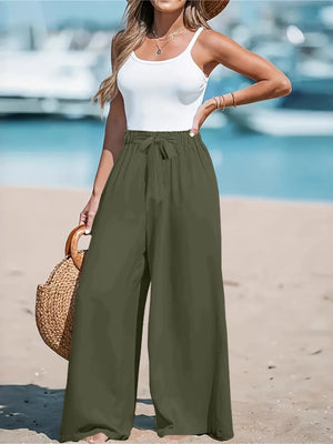 Woman in white top and green wide-leg pants standing on a beach with boats in the background.