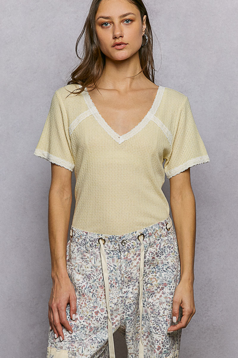 Woman wearing a light yellow top with lace trim and floral pants against a gray background