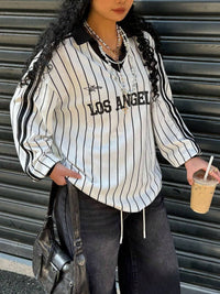 Person wearing a striped shirt with 'Los Angeles' text, holding a drink, standing against a metallic shutter.