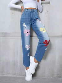 Blue jeans with floral patches worn by a person against a gray wall.