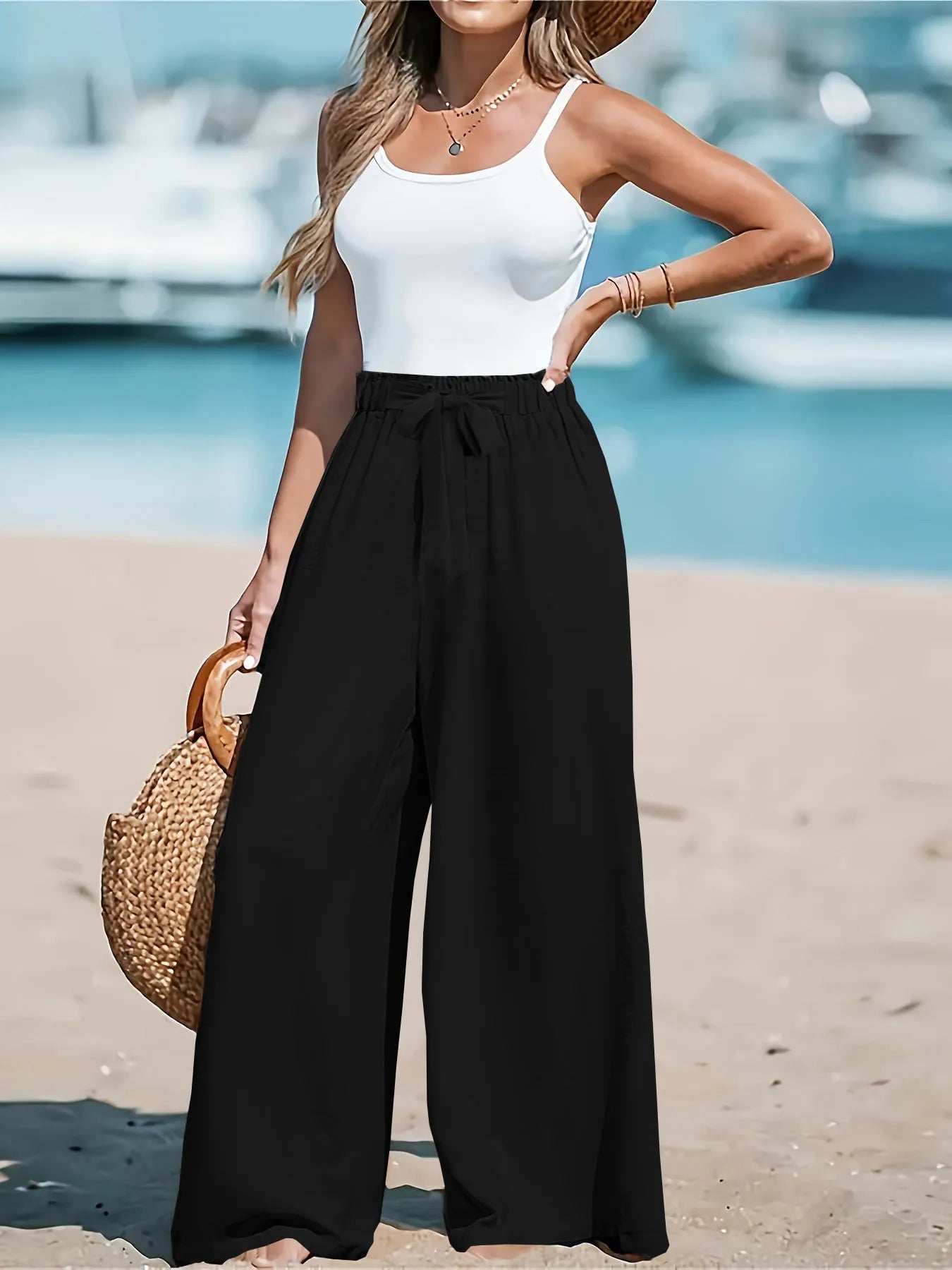 Woman in white top and black wide-leg pants standing on a beach with boats in the background.