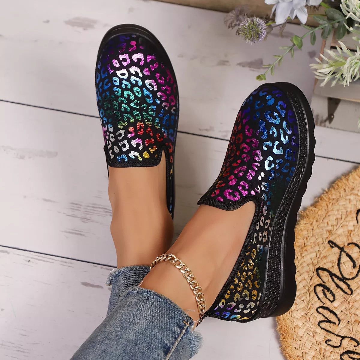 Multicolored leopard print shoes on a light wooden floor with a woven mat and flowers in the background.