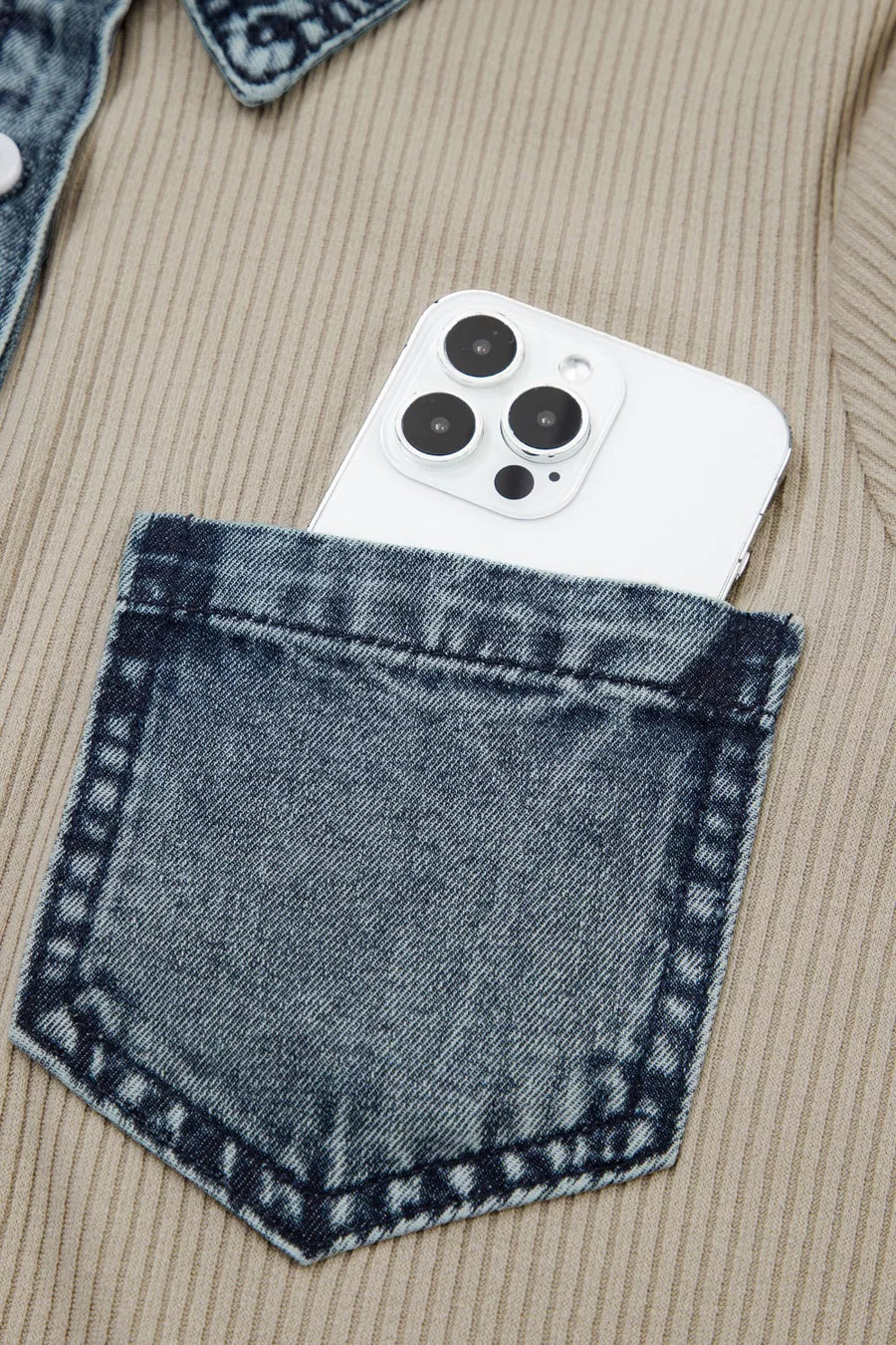 White phone with black camera details in a blue denim pocket of a beige jacket.
