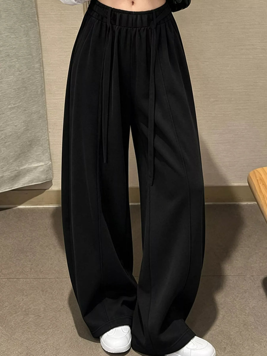 Black wide-leg drawstring pants worn by a model, paired with white sneakers, shown from the waist down in an indoor setting.