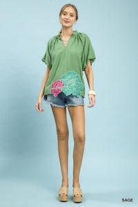 Woman wearing a green blouse with floral design and denim shorts on a light blue background