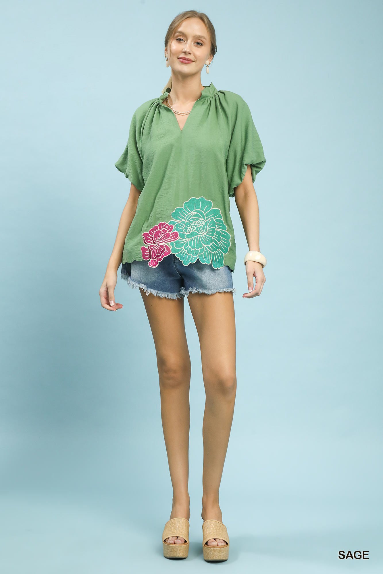 Woman wearing a green blouse with floral design and denim shorts on a light blue background
