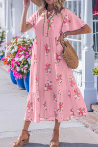 Woman wearing a pink floral dress standing outdoors with flowers and a building in the background.