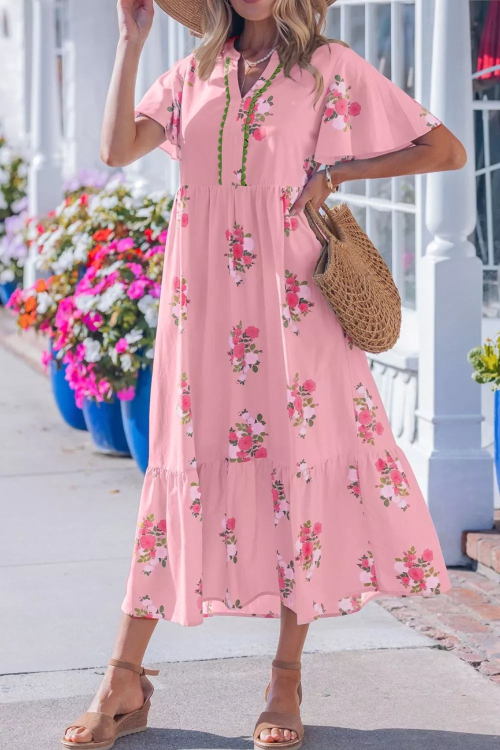 Woman wearing a pink floral dress standing outdoors with flowers and a building in the background.