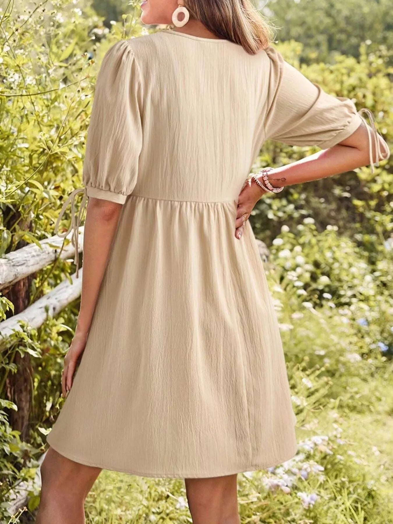 Woman wearing a beige dress in a natural setting with greenery and flowers.