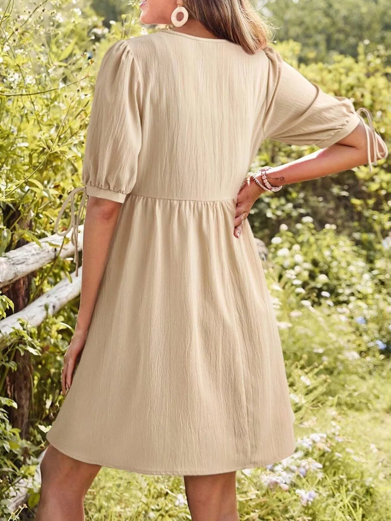 Woman wearing a beige dress in a natural setting with greenery and flowers.
