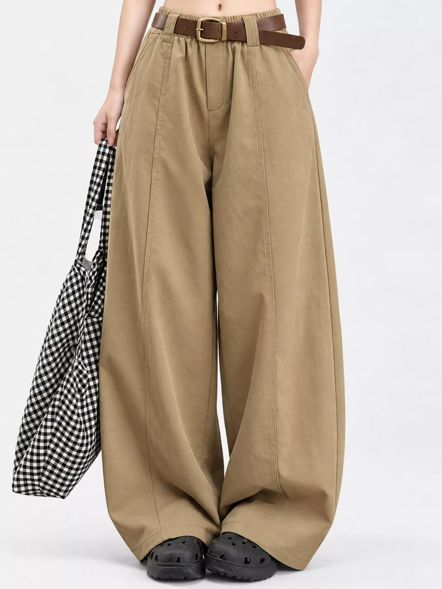 A model wearing high-waisted, wide-leg khaki pants with a brown belt, styled with black clogs and holding a black-and-white checkered tote bag against a plain white background.