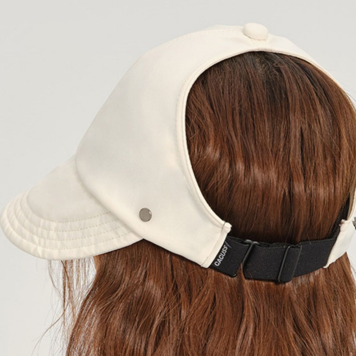 Beige cap with black strap worn by a person with brown hair on a light gray background