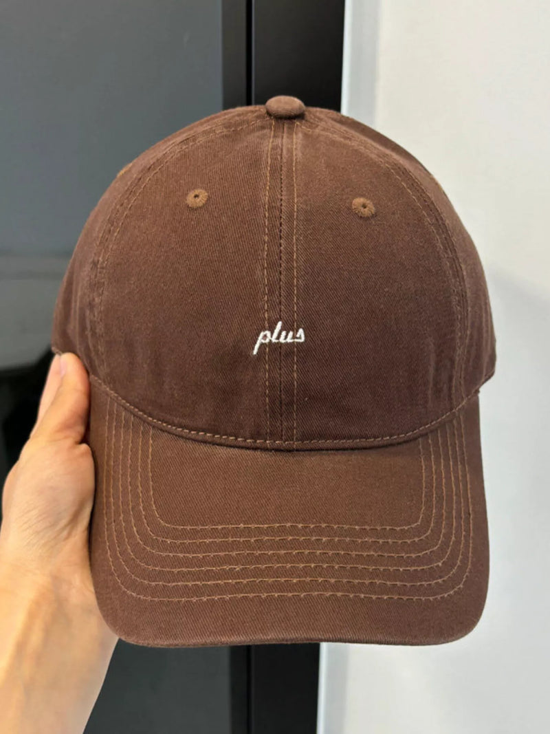 Brown cap with 'plus' logo held by a hand against a neutral background