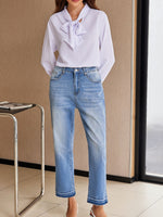 Person wearing a light purple blouse with a bow and blue jeans indoors.