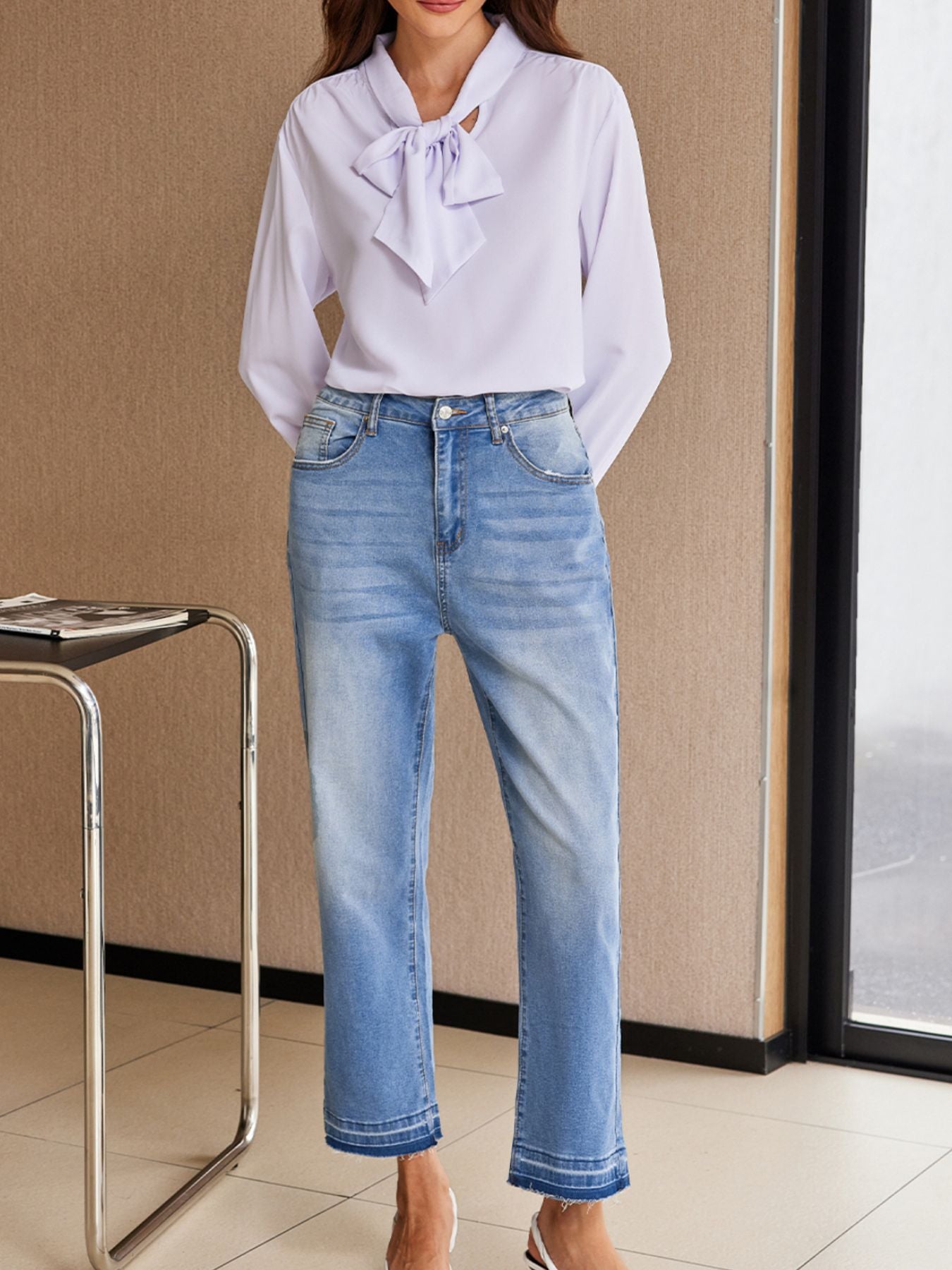 Person wearing a light purple blouse with a bow and blue jeans indoors.