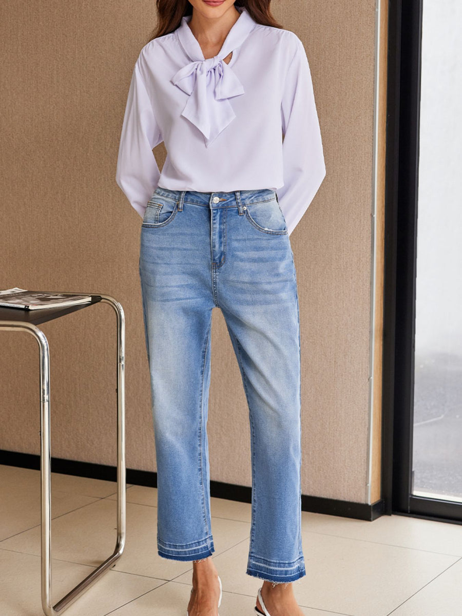 Person wearing a light purple blouse with a bow and blue jeans indoors.