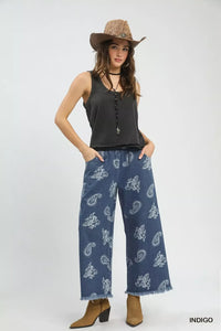 Woman wearing a black sleeveless top and blue paisley pants with a brown hat on a white background.